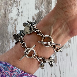 Puede incluir: Una pulsera plateada con cierre de palanca y varios dijes. La pulsera presenta un dise&ntilde;o de cadena gruesa con varios elementos decorativos, incluyendo peque&ntilde;os dijes circulares y una cuenta de cristal.