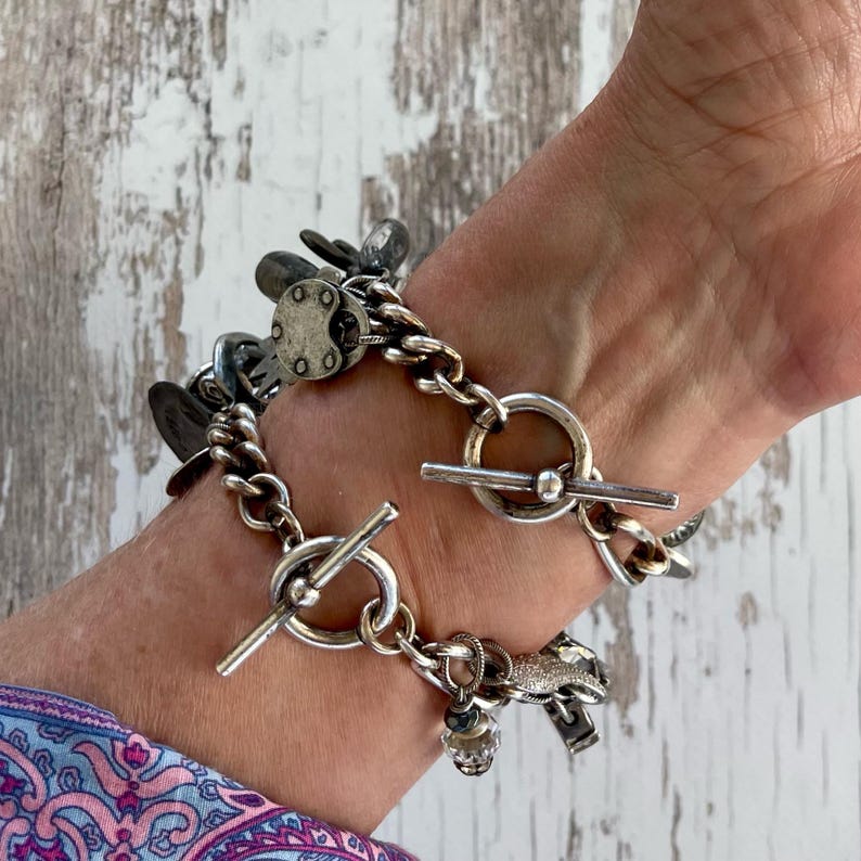 Puede incluir: Una pulsera plateada con cierre de palanca y varios dijes. La pulsera presenta un dise&ntilde;o de cadena gruesa con varios elementos decorativos, incluyendo peque&ntilde;os dijes circulares y una cuenta de cristal.