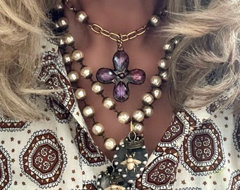 May include: A layered necklace with a large purple flower pendant and two smaller bee pendants. The necklace is made of gold chain and white pearls.