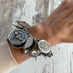Puede incluir: Una pulsera de plata con dijes de varios estilos antiguos, incluyendo monedas, medallones y un cristal brillante. La pulsera se lleva en la mu&ntilde;eca, mostrando los intrincados detalles y texturas de los dijes. Una pieza de joyer&iacute;a &uacute;nica.