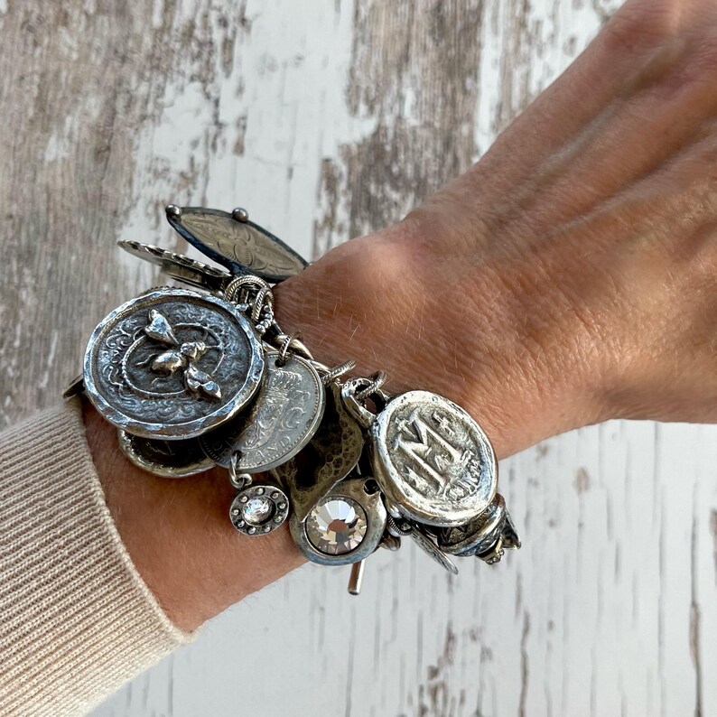 Puede incluir: Una pulsera de plata con dijes de varios estilos antiguos, incluyendo monedas, medallones y un cristal brillante. La pulsera se lleva en la mu&ntilde;eca, mostrando los intrincados detalles y texturas de los dijes. Una pieza de joyer&iacute;a &uacute;nica.