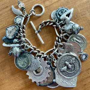 Puede incluir: Pulsera de plata con dijes variados y detallados, incluyendo monedas, engranajes y elementos decorativos. La pulsera presenta un cierre de palanca y una mezcla de texturas y formas, creando un accesorio &uacute;nico y ecl&eacute;ctico.