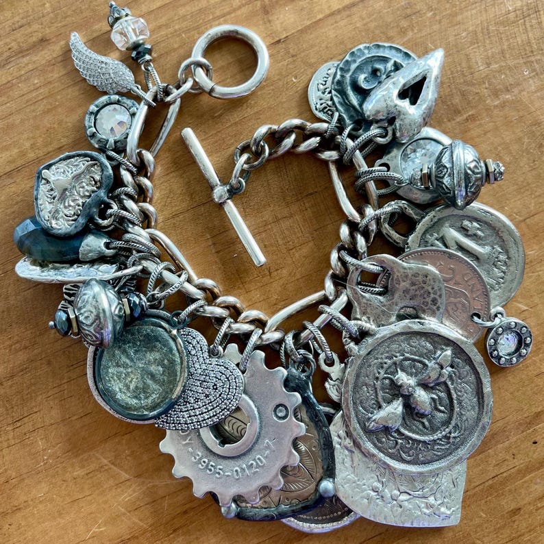 Puede incluir: Pulsera de plata con dijes variados y detallados, incluyendo monedas, engranajes y elementos decorativos. La pulsera presenta un cierre de palanca y una mezcla de texturas y formas, creando un accesorio &uacute;nico y ecl&eacute;ctico.