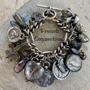 Puede incluir: Pulsera de plata con dijes variados, incluyendo monedas, un candado y una flor de lis. La pulsera tiene un cierre de palanca y un dise&ntilde;o de cadena. El texto "French Connection" es visible.