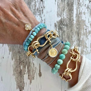 May include: A stack of bracelets featuring turquoise-colored beads, gold-toned metal chain links, and a fabric wrap. One bracelet includes a gold-toned clover charm with clear stones, and another features a round gold-toned coin pendant.