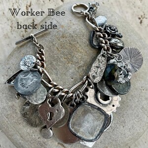 Puede incluir: Una pulsera de plata con varios dijes, incluyendo monedas, un candado en forma de coraz&oacute;n y una gema facetada. La pulsera tiene un cierre de palanca y se muestra sobre una superficie de piedra. El texto "Worker Bee back side" es visible.