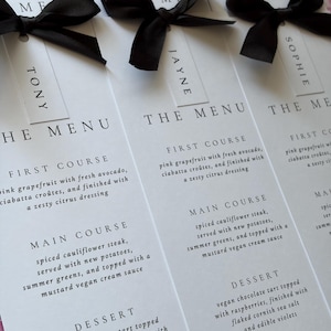 May include: White paper menu cards with black text and black satin ribbon bows. Each card features a name tag at the top, including Tony, Jayne, and Sophie. The menu lists a first course of pink grapefruit, a main course of spiced cauliflower steak, and a vegan chocolate tart dessert, dated 27.06.2026.