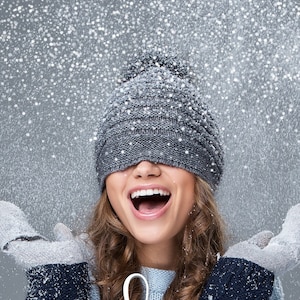May include: A woman wearing a gray knit beanie with a pom pom, smiling and laughing while snow falls around her. She is wearing a gray sweater and gloves.