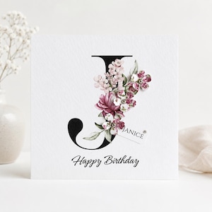 Puede incluir: Una tarjeta de cumplea&ntilde;os blanca con una letra "J" negra adornada con flores rosas y burdeos. La tarjeta incluye el nombre "Janice" en una peque&ntilde;a etiqueta y las palabras "Happy Birthday" en una elegante escritura.