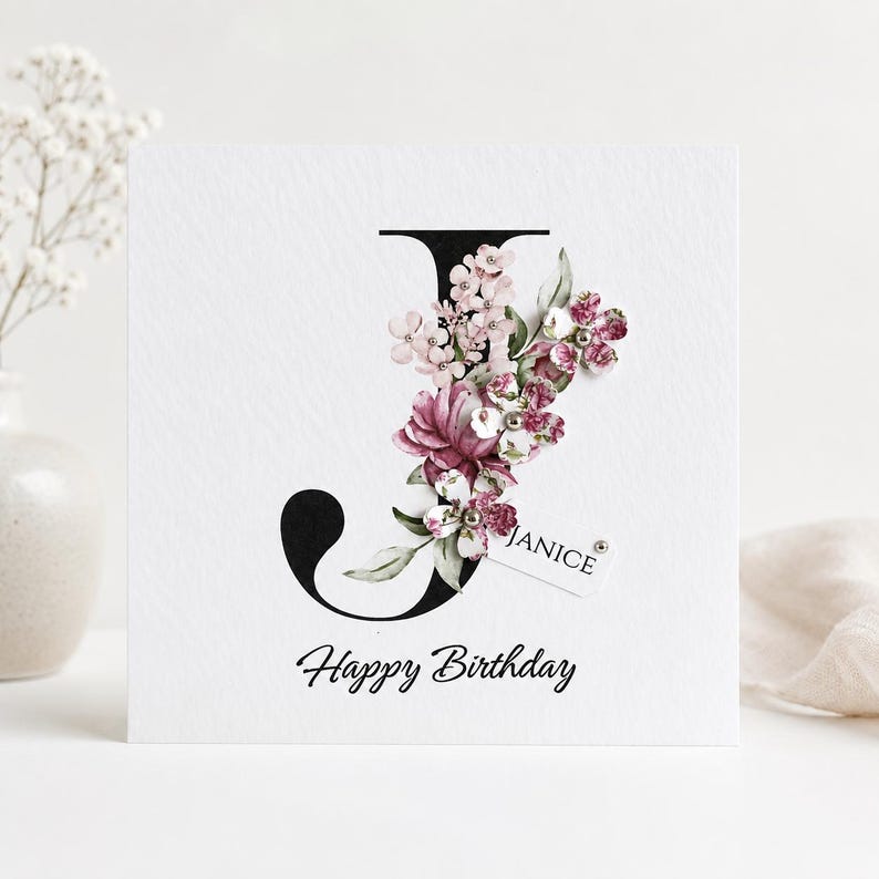 Puede incluir: Una tarjeta de cumplea&ntilde;os blanca con una letra "J" negra adornada con flores rosas y burdeos. La tarjeta incluye el nombre "Janice" en una peque&ntilde;a etiqueta y las palabras "Happy Birthday" en una elegante escritura.