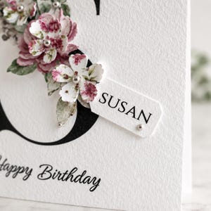 Puede incluir: Una tarjeta de cumplea&ntilde;os blanca con la inscripci&oacute;n manuscrita "Happy Birthday". La tarjeta presenta una gran letra negra y est&aacute; adornada con flores de papel en tonos rosas y blancos, con el nombre "SUSAN" en una etiqueta.