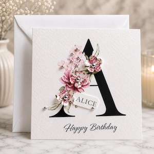 Puede incluir: Una tarjeta de felicitaci&oacute;n blanca con una gran letra "A" negra adornada con flores rosas y plateadas. La tarjeta incluye el nombre "ALICE" y las palabras "Happy Birthday" en escritura negra. Hay un sobre blanco detr&aacute;s de la tarjeta.