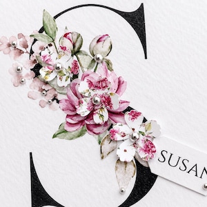 Puede incluir: Una letra "S" decorativa con un arreglo floral. Las flores son rosas y blancas con detalles plateados. La palabra "SUSAN" est&aacute; en una peque&ntilde;a etiqueta blanca. El fondo es blanco con una letra negra.