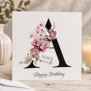 Puede incluir: Tarjeta de felicitaci&oacute;n blanca con una gran letra 'A' negra adornada con flores rosas y blancas. La tarjeta tiene el nombre 'Alice' en una peque&ntilde;a etiqueta y la frase 'Happy Birthday' en la parte inferior.