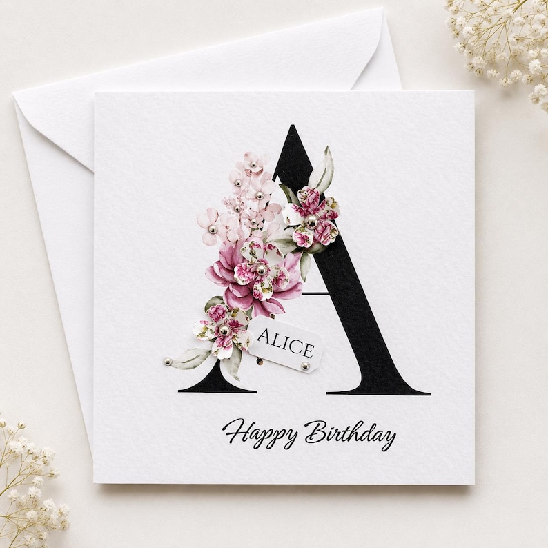 Puede incluir: Tarjeta de felicitaci&oacute;n blanca con una gran letra "A" negra adornada con flores rosas y blancas. La tarjeta incluye el nombre "Alice" en una peque&ntilde;a etiqueta y la frase "Happy Birthday" en cursiva.