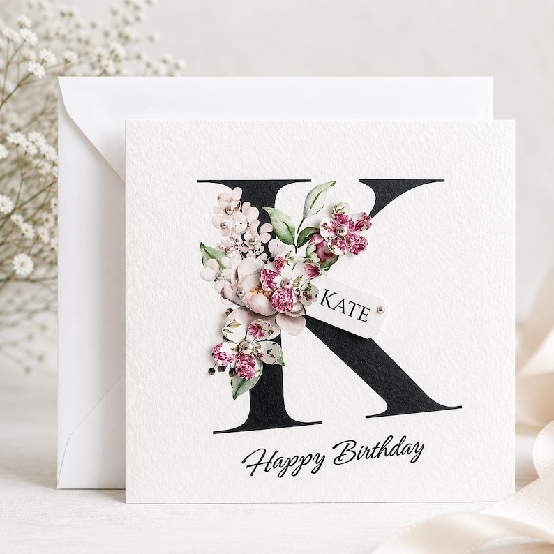 Puede incluir: Una tarjeta de felicitaci&oacute;n blanca cuadrada con una gran letra negra "K" adornada con arreglos florales rosas y blancos. El nombre "KATE" est&aacute; en una peque&ntilde;a etiqueta. Las palabras "Happy Birthday" est&aacute;n escritas debajo en cursiva.
