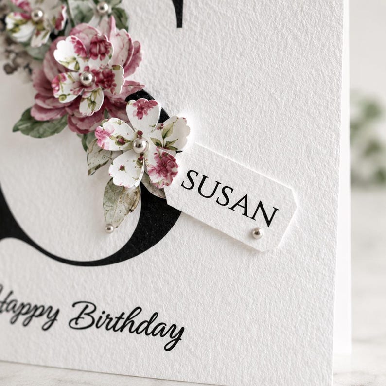 Puede incluir: Una tarjeta de cumplea&ntilde;os blanca con la inscripci&oacute;n manuscrita "Happy Birthday". La tarjeta presenta una gran letra negra y est&aacute; adornada con flores de papel en tonos rosas y blancos, con el nombre "SUSAN" en una etiqueta.