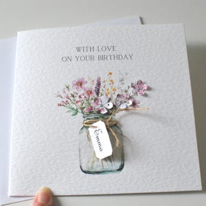 May include: A birthday card with the text "WITH LOVE ON YOUR BIRTHDAY". The card features a watercolor illustration of a mason jar filled with pink and purple flowers. A small tag with the name "Emma" is attached.
