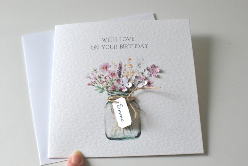 May include: A birthday card with the text "WITH LOVE ON YOUR BIRTHDAY". The card features a watercolor illustration of a mason jar filled with pink and purple flowers. A small tag with the name "Emma" is attached.