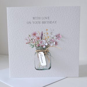 May include: A birthday card with a watercolor illustration. The card features a glass jar filled with pink and purple flowers tied with twine. The text "WITH LOVE ON YOUR BIRTHDAY" is printed at the top. A tag with the name "Emma" is attached.