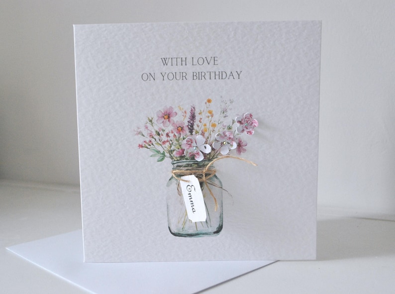 May include: A birthday card with a watercolor illustration. The card features a glass jar filled with pink and purple flowers tied with twine. The text "WITH LOVE ON YOUR BIRTHDAY" is printed at the top. A tag with the name "Emma" is attached.