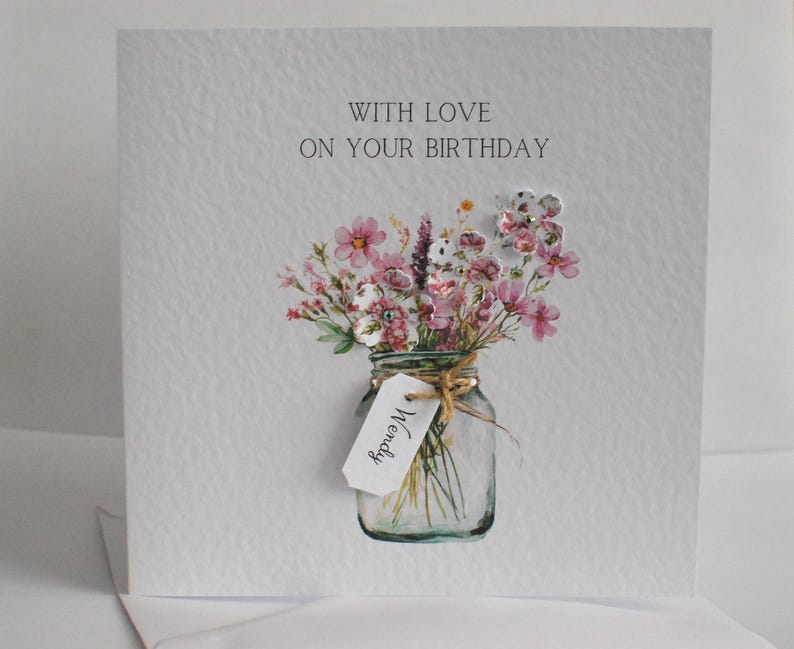 May include: A handmade birthday card with a watercolor illustration of a mason jar filled with pink and white flowers. The card reads "WITH LOVE ON YOUR BIRTHDAY" and has a small tag that says "Wendy".