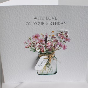 May include: A white birthday card with the text "WITH LOVE ON YOUR BIRTHDAY". The card features a watercolor illustration of a mason jar filled with pink and white flowers. A tag with the name "Wendy" is tied to the jar.