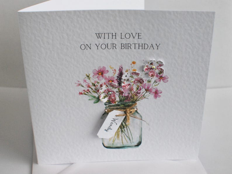 May include: A white birthday card with the text "WITH LOVE ON YOUR BIRTHDAY". The card features a watercolor illustration of a mason jar filled with pink and white flowers. A tag with the name "Wendy" is tied to the jar.