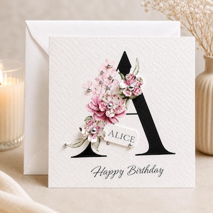 Puede incluir: Tarjeta de felicitaci&oacute;n blanca con una gran letra negra "A" adornada con flores rosas y blancas. La tarjeta incluye el nombre "ALICE" en una peque&ntilde;a etiqueta y las palabras "Happy Birthday" en cursiva.