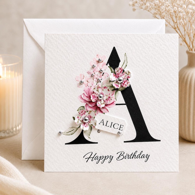 Puede incluir: Tarjeta de felicitaci&oacute;n blanca con una gran letra negra "A" adornada con flores rosas y blancas. La tarjeta incluye el nombre "ALICE" en una peque&ntilde;a etiqueta y las palabras "Happy Birthday" en cursiva.