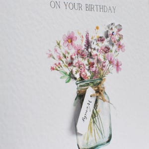 May include: A birthday card with the text "ON YOUR BIRTHDAY" at the top. The card features a watercolor illustration of a mason jar filled with pink and purple flowers. A tag with the name "Wendy" hangs from the jar.