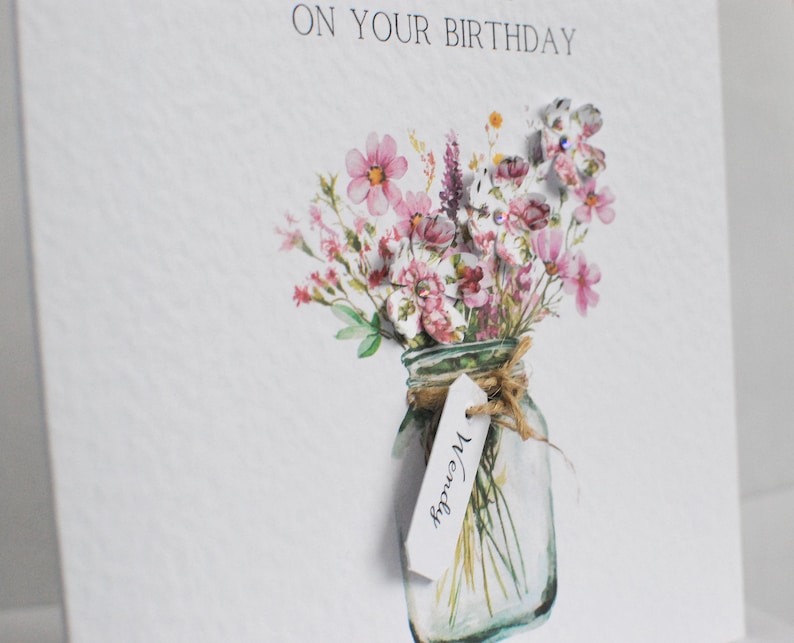 May include: A birthday card with the text "ON YOUR BIRTHDAY" at the top. The card features a watercolor illustration of a mason jar filled with pink and purple flowers. A tag with the name "Wendy" hangs from the jar.