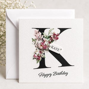 Puede incluir: Una tarjeta de cumplea&ntilde;os blanca con una gran letra negra "K" adornada con flores rosas y burdeos. El nombre "Kate" est&aacute; en una peque&ntilde;a etiqueta. Las palabras "Happy Birthday" est&aacute;n escritas en la parte inferior. Se ve un sobre.