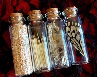 May include: Four small glass bottles with cork tops, each containing a different natural item. The bottles are arranged in a row on a black and red lace fabric. The items in the bottles are: a pile of small white and beige granules, a bundle of thin, light brown sticks, a dried, light brown snake skin, and a white and black butterfly wing.