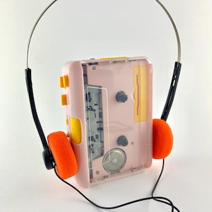 May include: A vintage pink and yellow cassette player with orange ear pads and a silver headband. The clear plastic case reveals the internal components. Black wires connect the headphones to the player, creating a retro aesthetic.