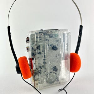 May include: A vintage clear plastic cassette player with a silver headband and black earphone arms. The headphones have bright orange foam ear pads. The player has visible internal components and a black cord.