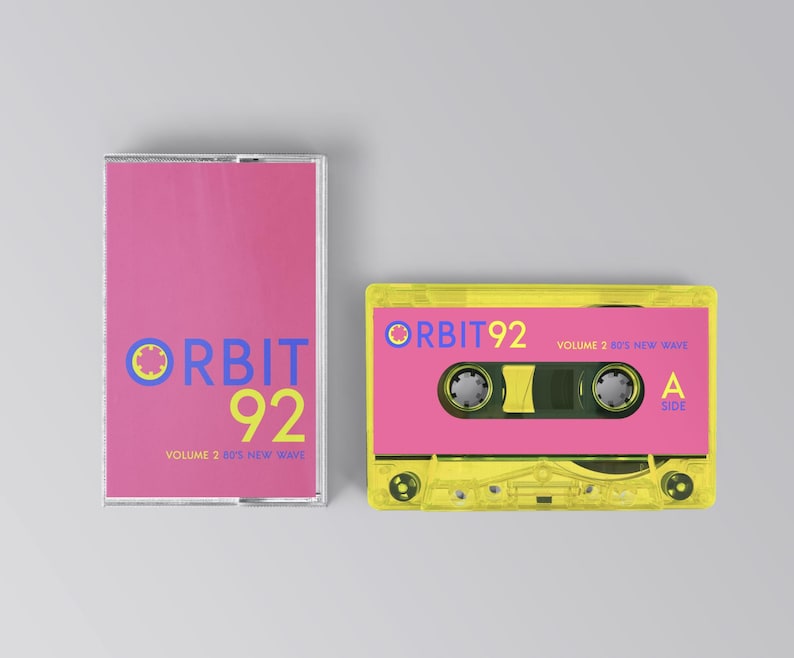 May include: A yellow cassette tape with a pink label. The label has the text "ORBIT 92" in blue and yellow, and "VOLUME 2 80'S NEW WAVE" in smaller white text. The cassette tape has a clear plastic case and a black tape spool.