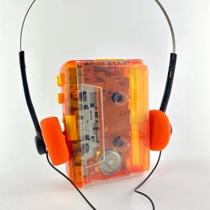 May include: A vintage orange and clear cassette player with matching orange foam ear pads. The headphones have a silver band and black accents. The player's internal components are visible through the transparent casing.