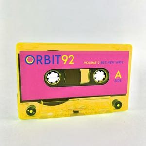 May include: A yellow cassette tape with the text "ORBIT 92 VOLUME 2 80'S NEW WAVE" on the label. The label is pink and has a small blue circle with a white dot in the center. The tape is labeled "A SIDE" on the right side.