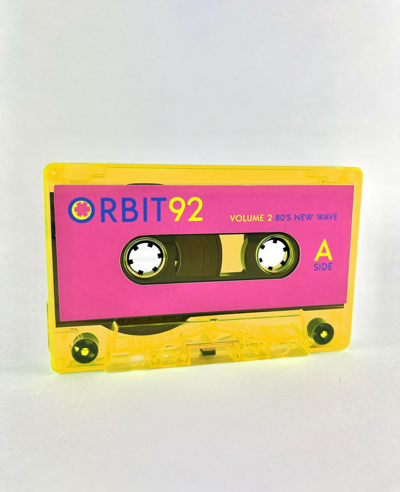 May include: A yellow cassette tape with the text "ORBIT 92 VOLUME 2 80'S NEW WAVE" on the label. The label is pink and has a small blue circle with a white dot in the center. The tape is labeled "A SIDE" on the right side.