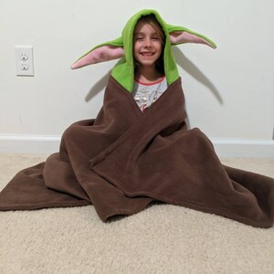 May include: A child wearing a green and pink hooded blanket with large ears, resembling a popular fictional character. The blanket is brown and made of fleece.
