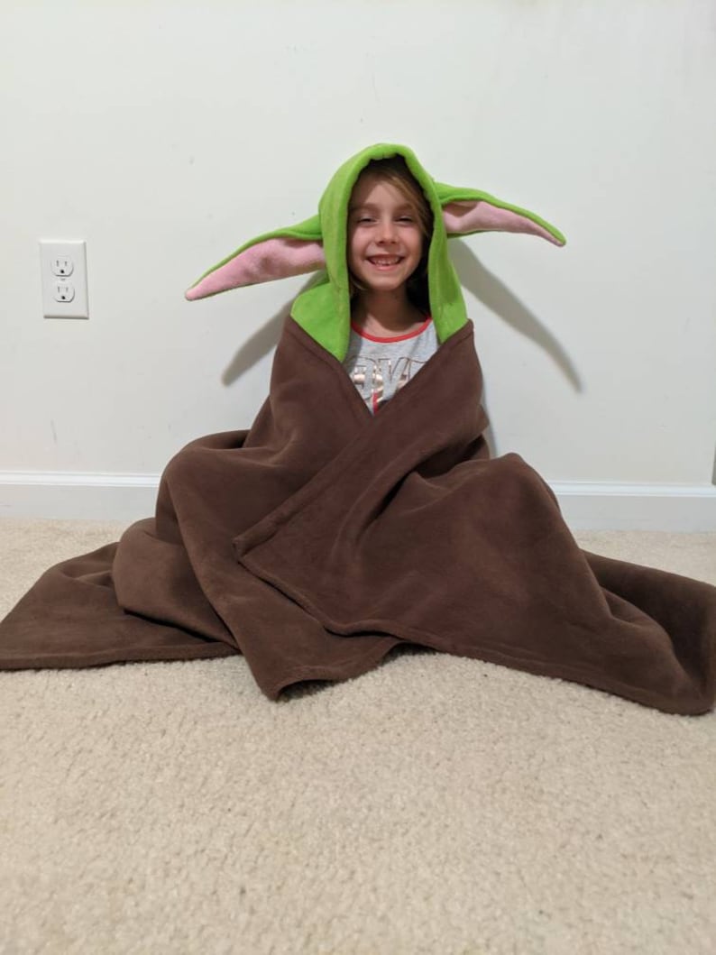 May include: A child wearing a green and pink hooded blanket with large ears, resembling a popular fictional character. The blanket is brown and made of fleece.