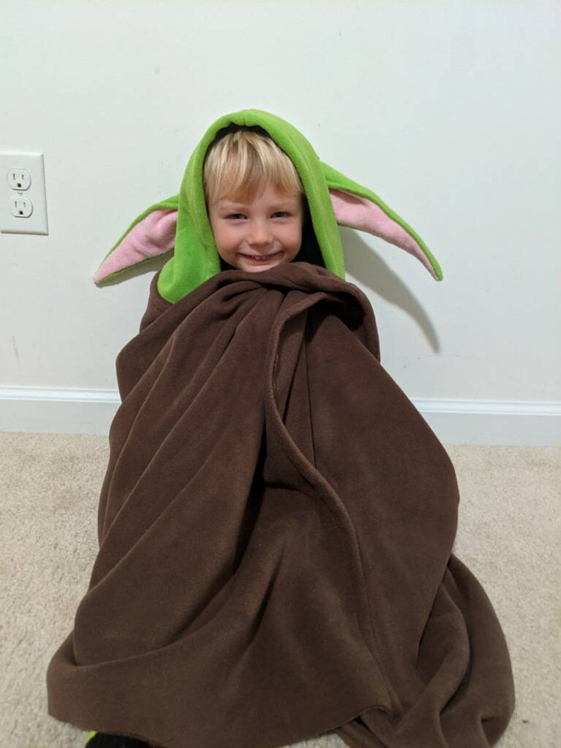 May include: A young child wearing a green and pink hooded blanket with large ears. The child is smiling and looking at the camera. The blanket is brown and has a soft, fuzzy texture.