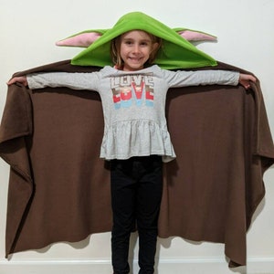 May include: A child wearing a green hooded blanket with pink ears and a grey shirt with the word "LOVE" printed on it. The blanket is brown on the inside and has a long cape.