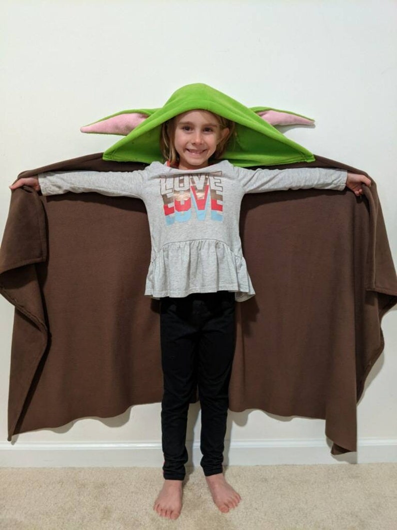 May include: A child wearing a green hooded blanket with pink ears and a grey shirt with the word "LOVE" printed on it. The blanket is brown on the inside and has a long cape.
