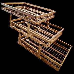 May include: A three-tiered wooden display rack with a natural finish. Each tier is a rectangular basket with a slatted base and a decorative railing. The tiers are connected by hinged supports, allowing them to be angled.