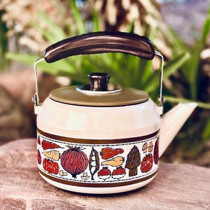 May include: Cream-colored vintage tea kettle with a brown handle and lid. The kettle features a decorative band of colorful vegetables, including tomatoes, onions, and carrots. The spout and handle are silver.