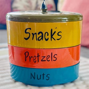 May include: A three-tiered, cylindrical snack container with a pointed finial. The top section is yellow with "Snacks" in black script, the middle is orange with "Pretzels", and the bottom is blue with "Nuts". The lid and top rim are olive green.