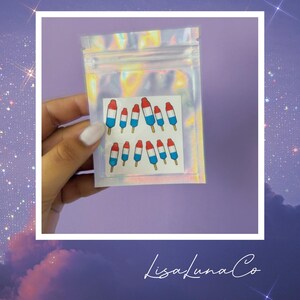 May include: A clear, iridescent bag containing a sheet of popsicle-shaped temporary tattoos. The popsicles are red, white, and blue, with wooden sticks. The bag is held by a hand with a white nail polish. The text "LisaLunaCo" is at the bottom.