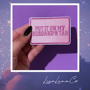 May include: Pink embroidered patch with white stitching. The patch reads "Put it on my husband's tab".